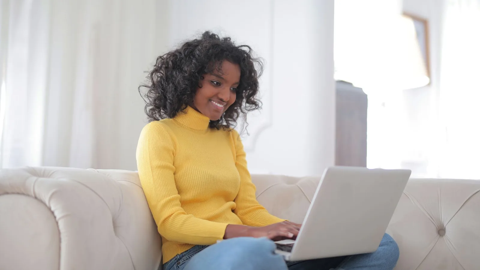 Positive young ethnic lady in casual yellow sweater and jeans sitting on comfortable couch and working on laptop while spending time at home