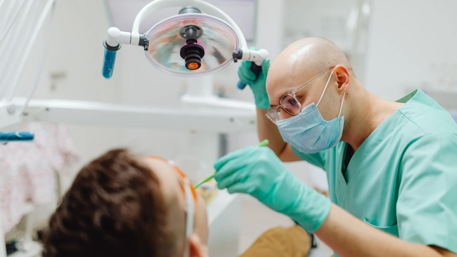 A dentist in green scrubs treating a patient under dental light in a clinic.
