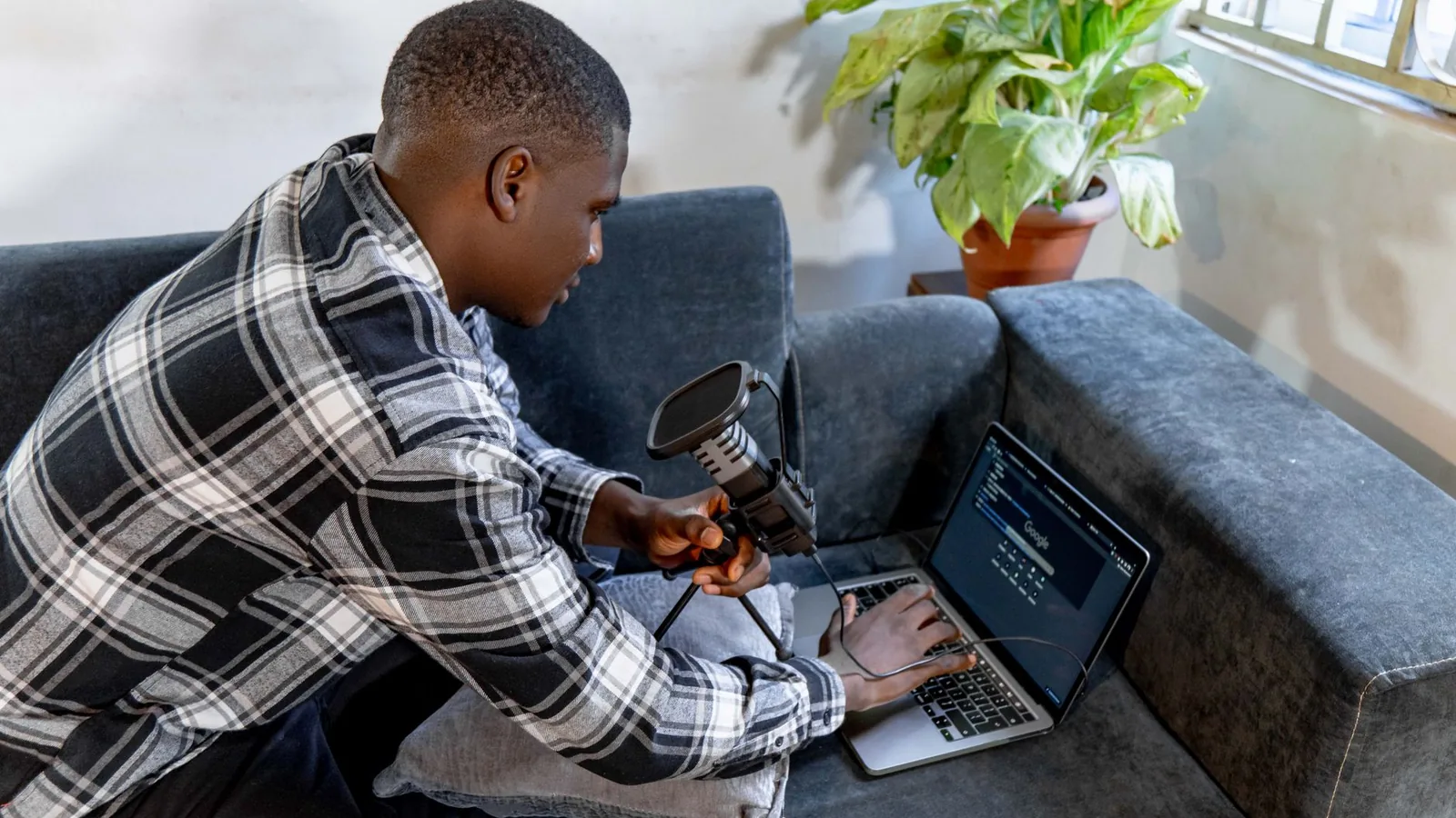 Young man sitting on a sofa using a laptop with a microphone, indoor setting.