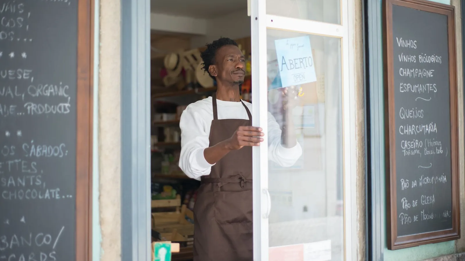 A man in an apron opens a grocery store door in Portugal, ready for business.