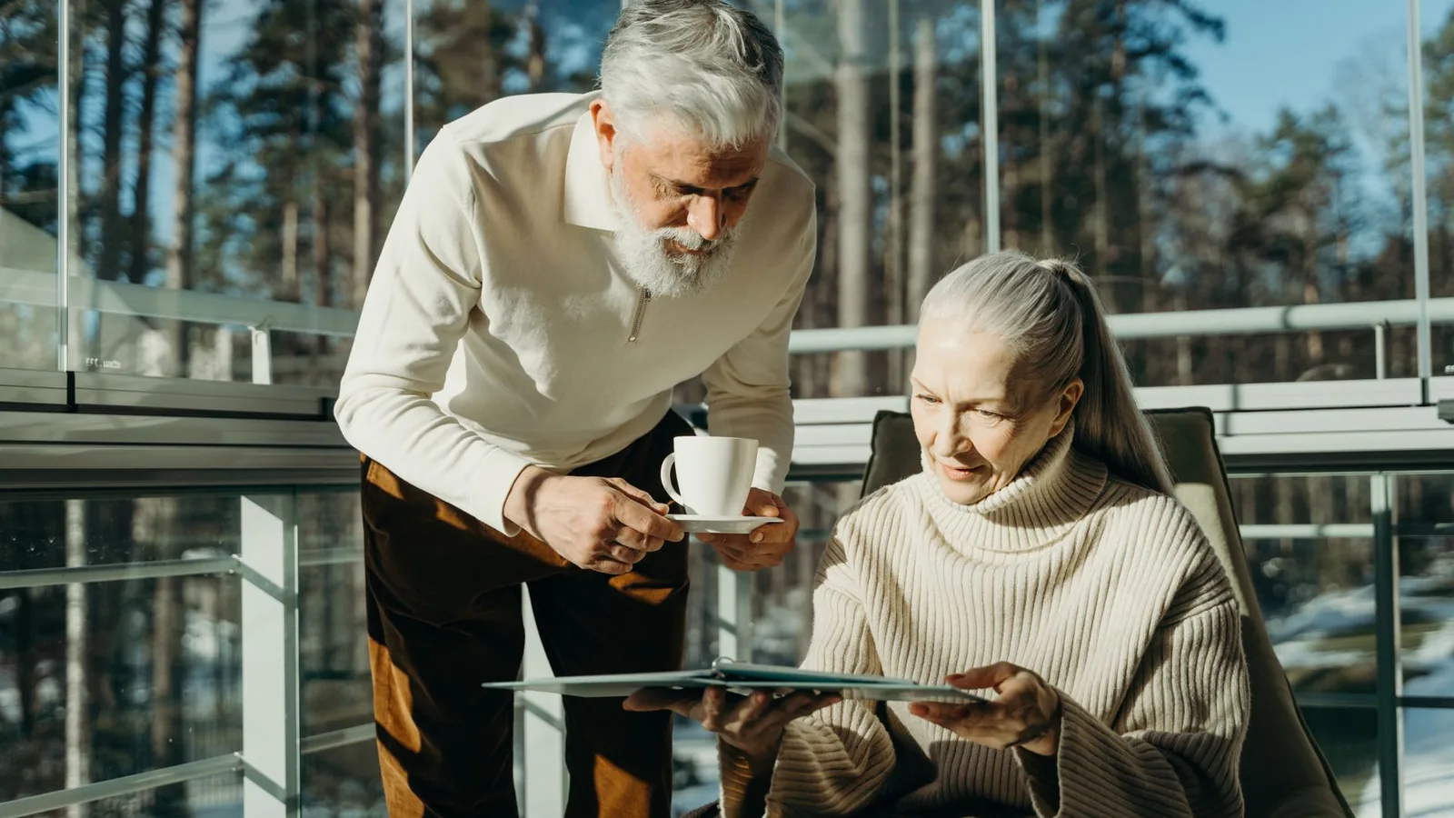 Senior couple enjoying coffee and reading in a sunlit room with large windows.
