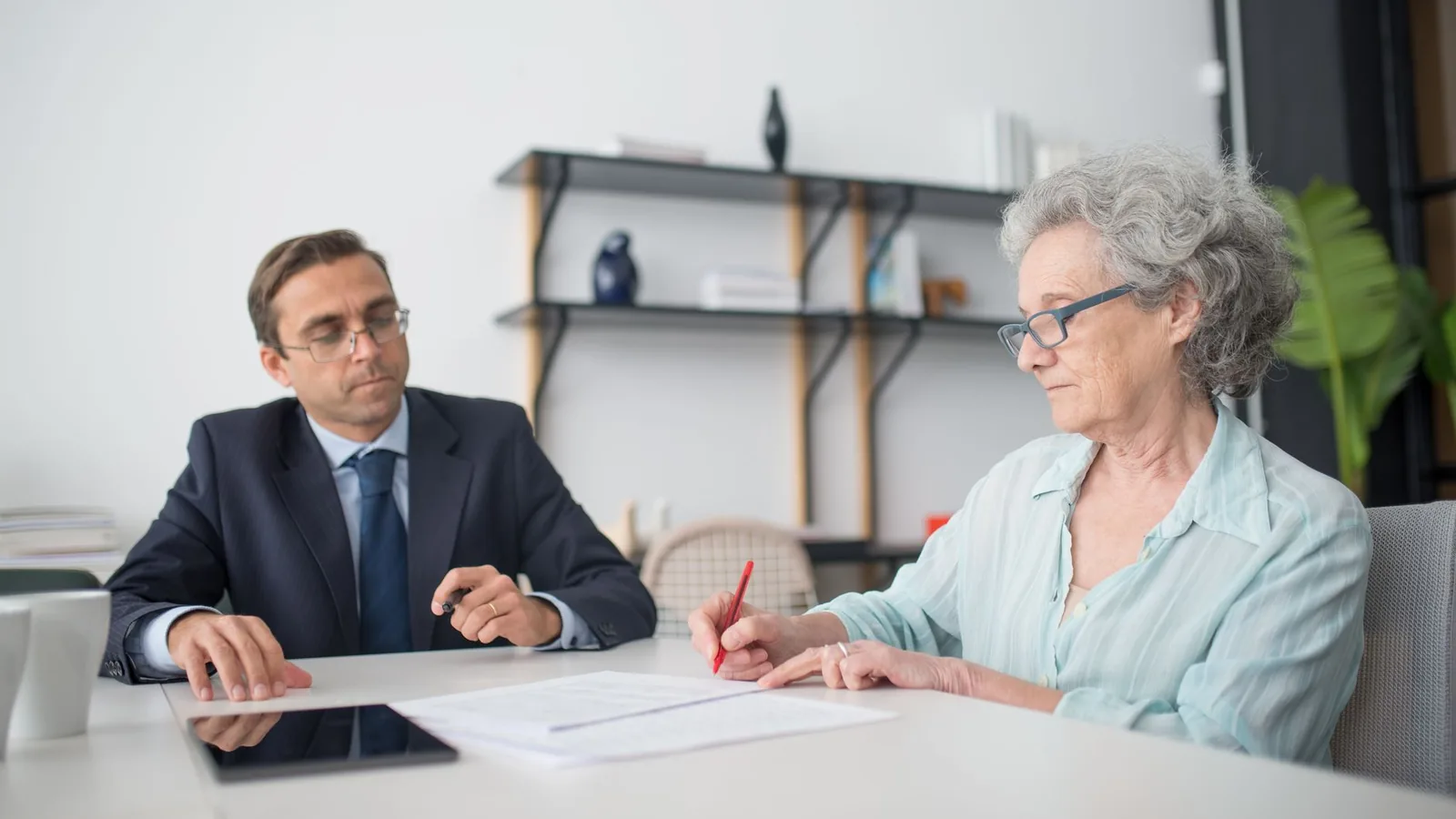 Elderly woman signing paperwork in modern office with consultant.