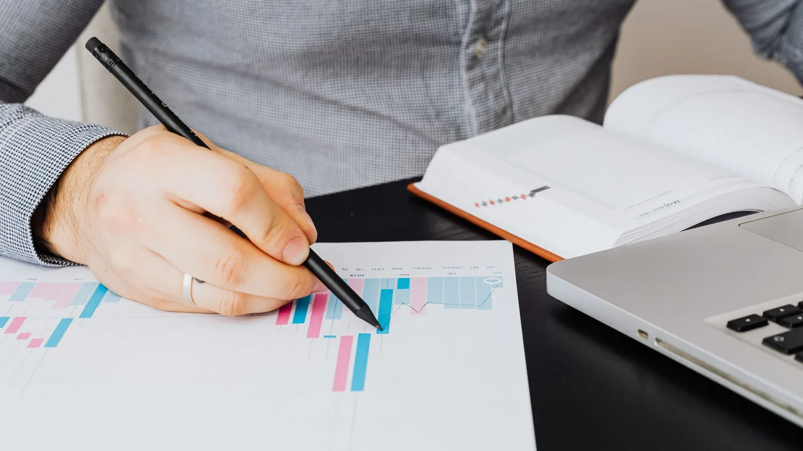 A business analyst examines a graph on paper with a laptop and notebook indoors.