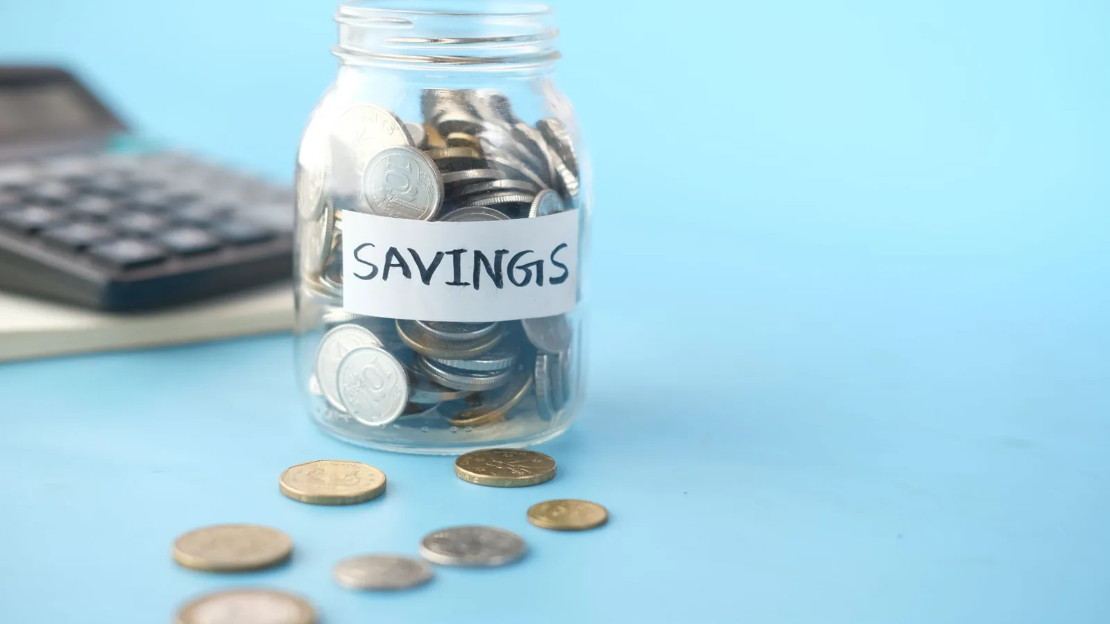 Glass jar labeled 'Savings' filled with coins, beside a calculator on a blue background.