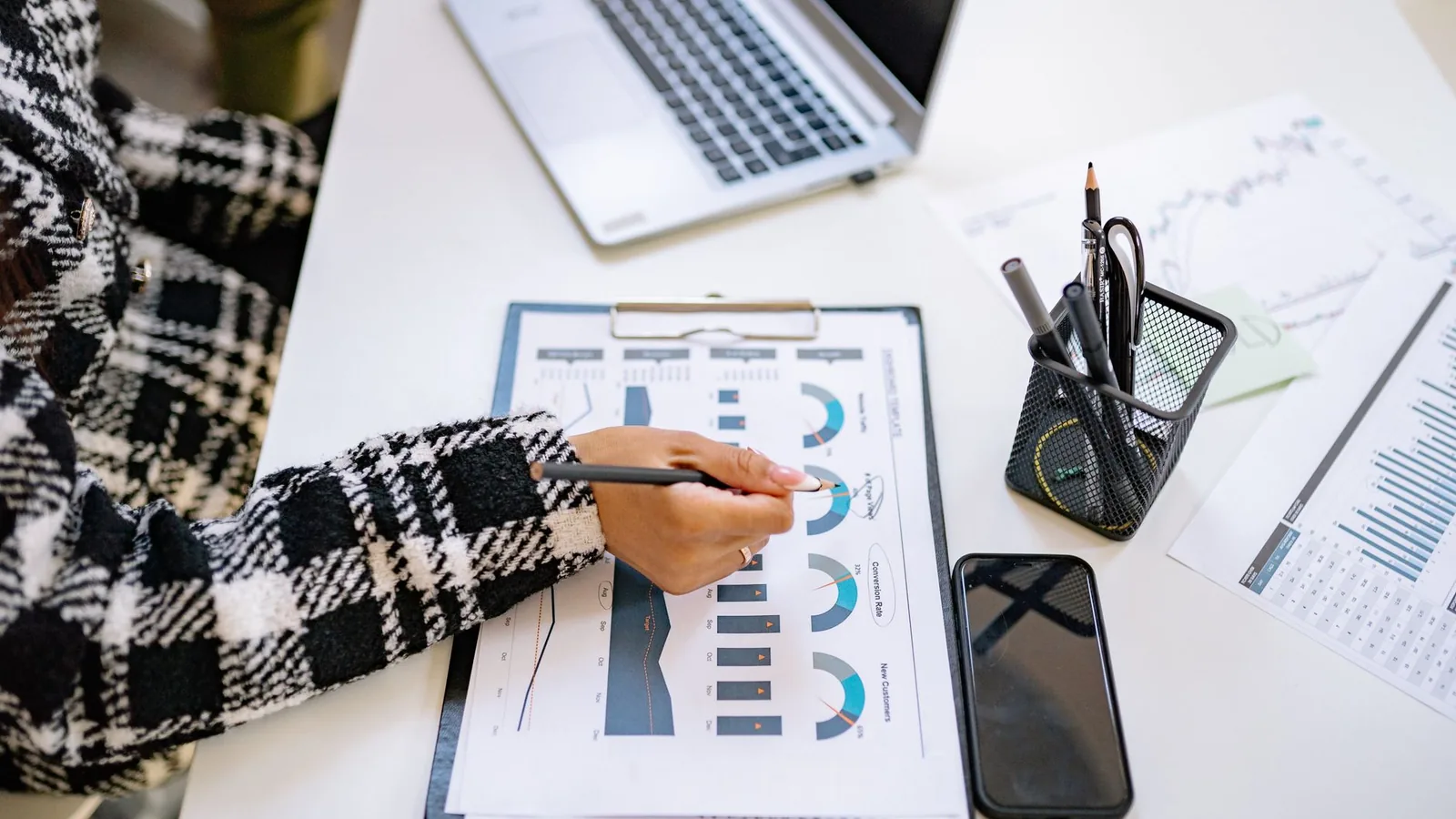 An office setting with a person analyzing charts and graphs at a desk with a laptop and smartphone.