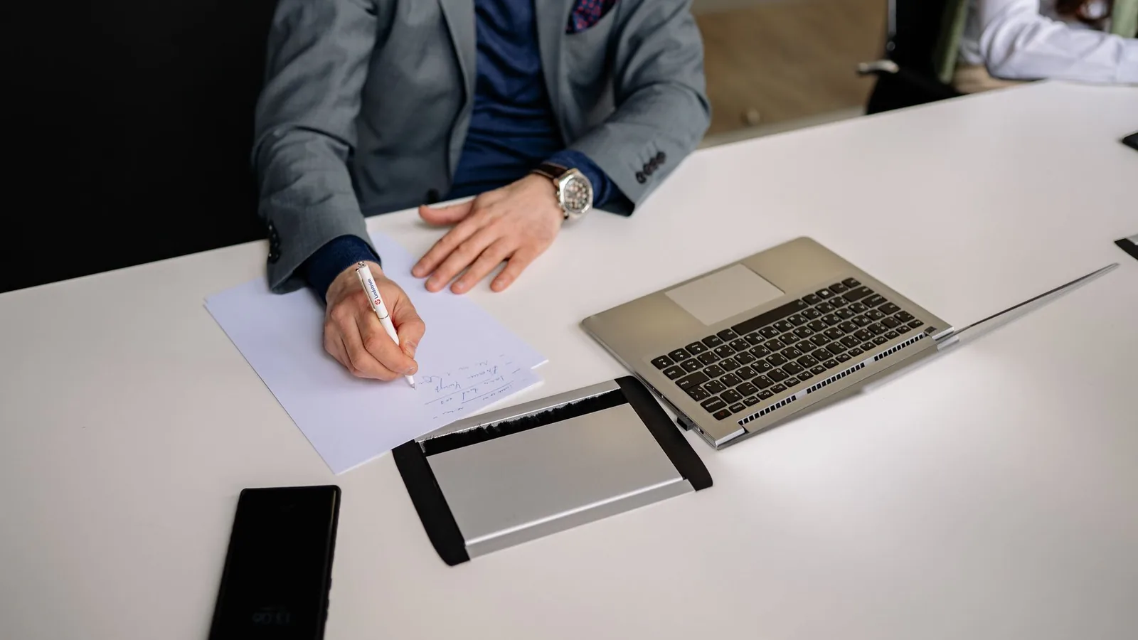 Professional businessman writing notes in a modern office environment with laptop and smartphone.