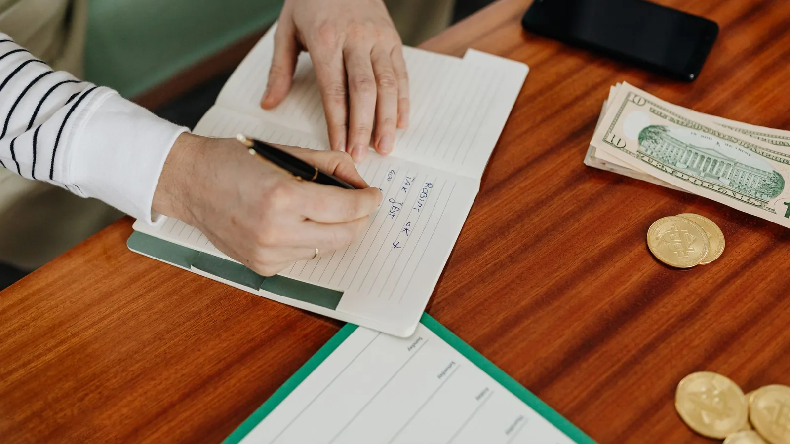 Person writing in a notebook on a wooden table with cash, coins, and a phone nearby.