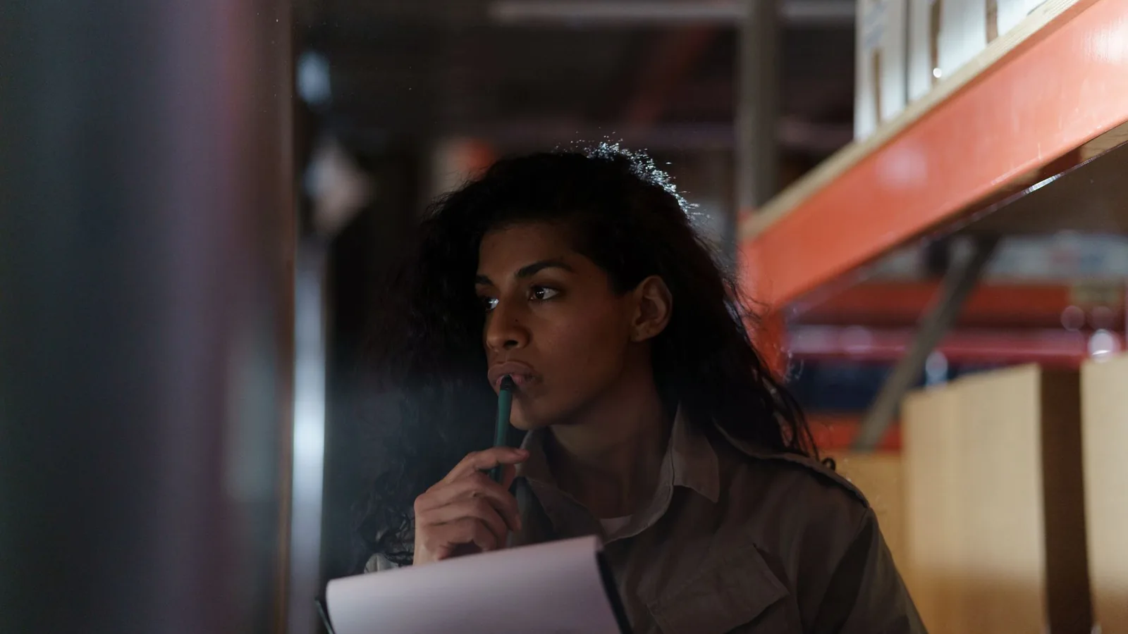 A woman in a warehouse contemplatively checking inventory under low light conditions.
