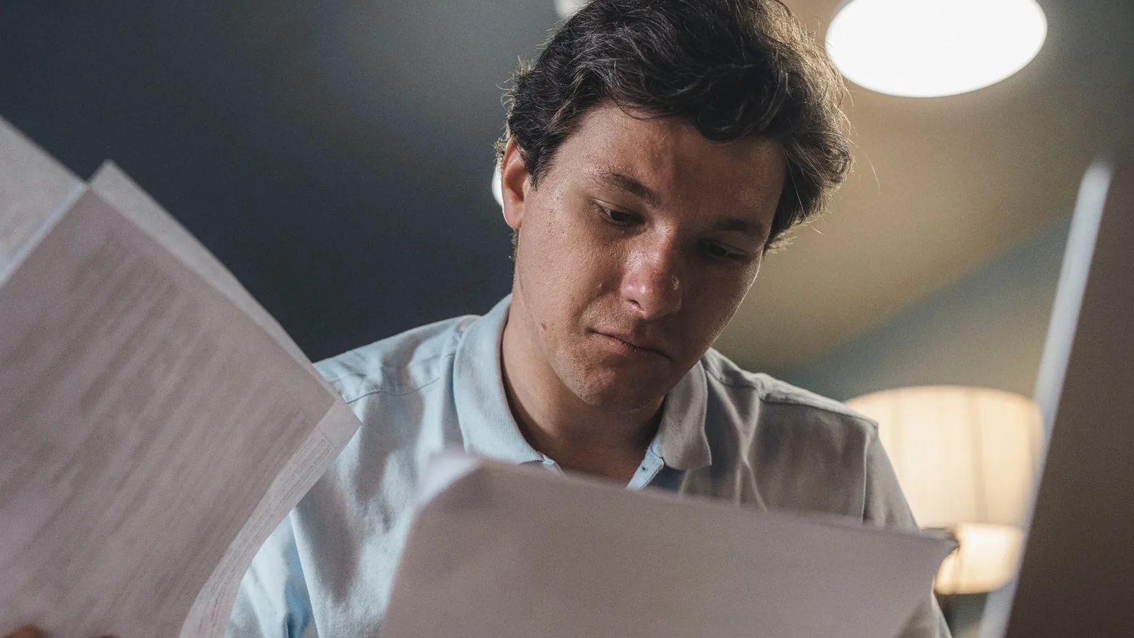 Serious man wearing a light blue shirt intently reading printed papers indoors under warm lighting.