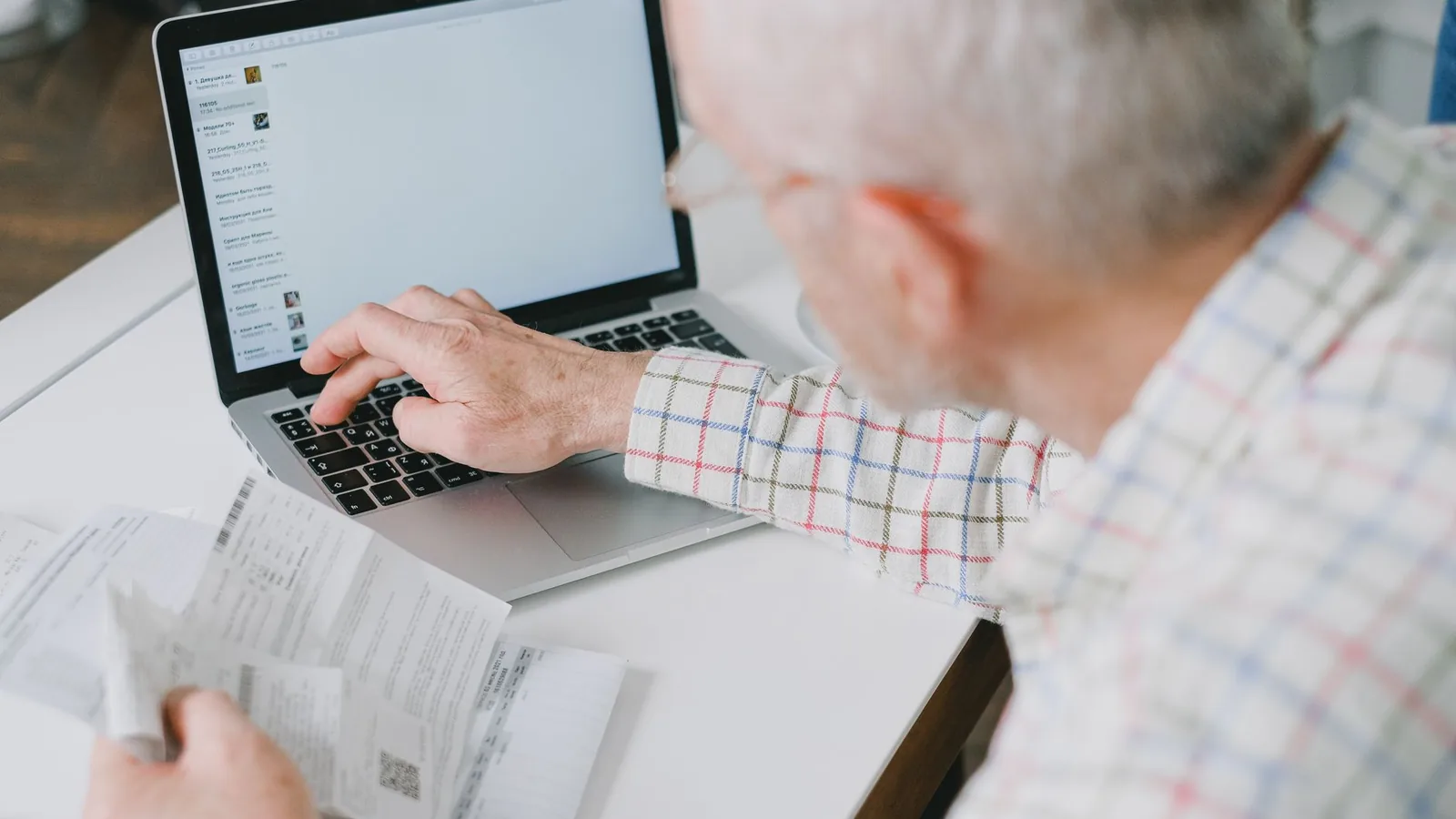 Elderly man at desk handling finances online with documents and laptop.