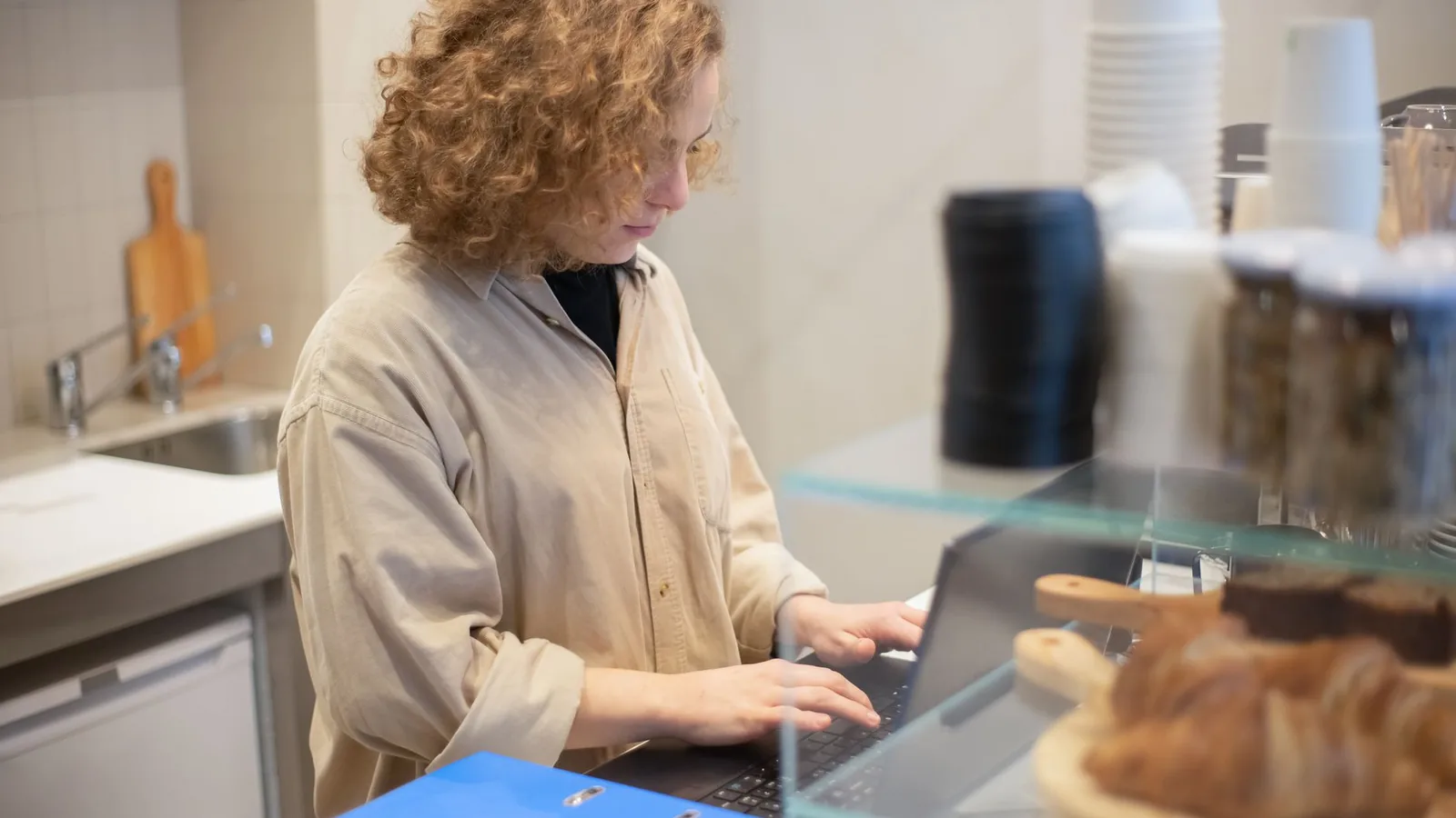 A young woman uses a laptop behind a café counter with pastries in foreground.
